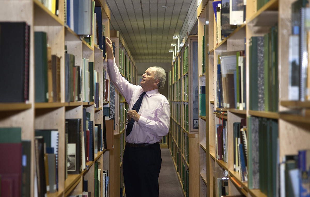 A man dressed in a shirt and tie selects a book from a shelf in a library. He is flanked by bookshelves on either side which contain many books of a variety of colours and sizes. 