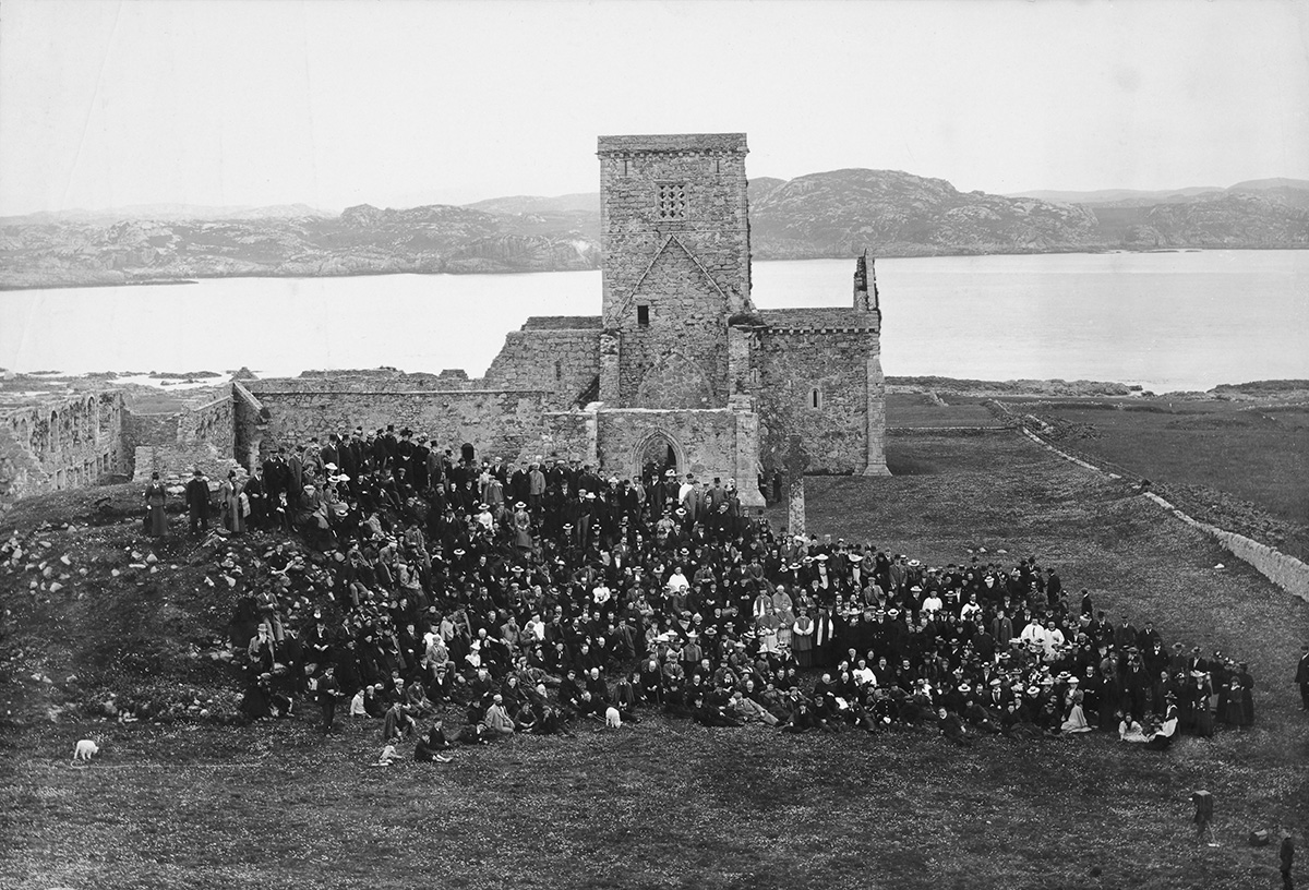 A large crowd is gathered in front of Iona Abbey. The photo is black and white and the sea and Isle of Mull are in the background.