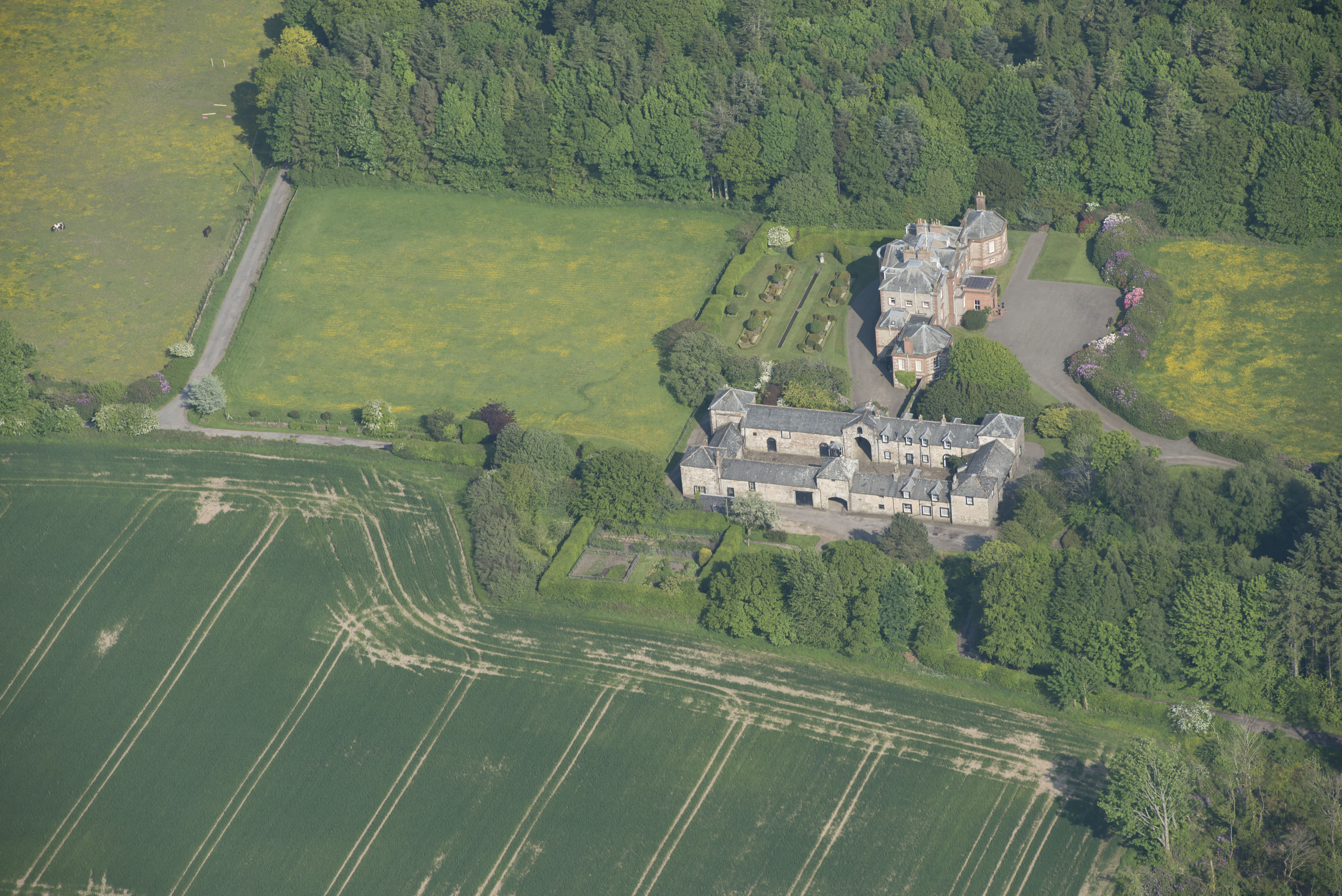 An aerial shot of a country estate. There is a house, stables and outbuildings. Next to the house there is a well-kept garden. Farmland and trees surround the estate. 