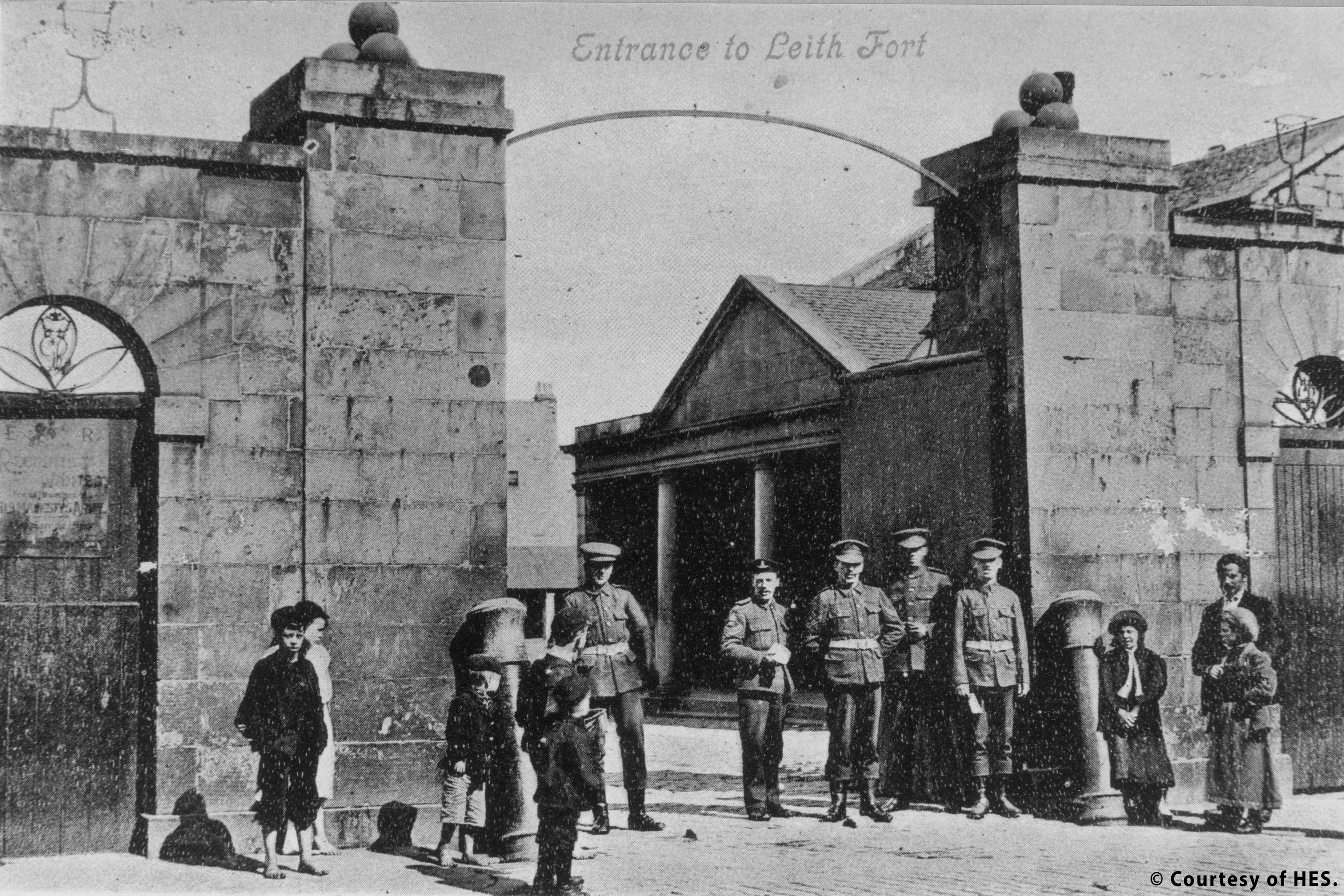 An archive image of the entrance to Leith fort. A large stone gateway is open showing a glimpse of fort buildings. Five soldiers in uniform are standing in the gateway. A number of local children are standing nearby. 