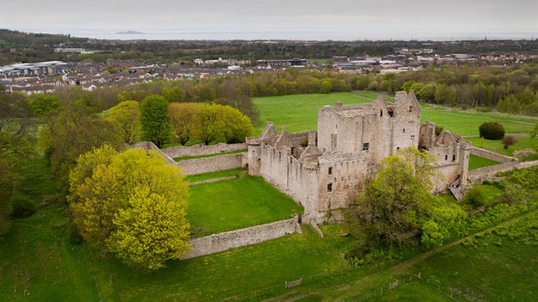 A view of Craigmillar Castle, venue for Spotlight on Mary. The city of Edinburgh and the Firth of Forth can be seen in the background. The castle is surrounded by green fields and trees and a walled garden is evident. 