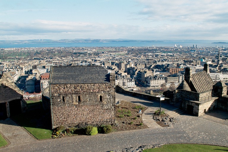 A small chapel sitting within the walls of Edinburgh Castle. The city of Edinburgh and the Firth of Forth can be seen in the background. 