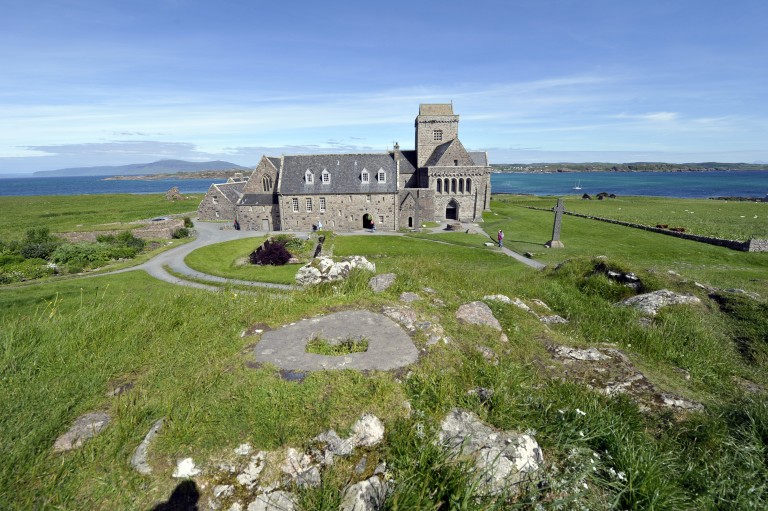 A general view of Iona Abbey with the sea in the background. A visitor and a stone cross can be seen to the right of the building. 