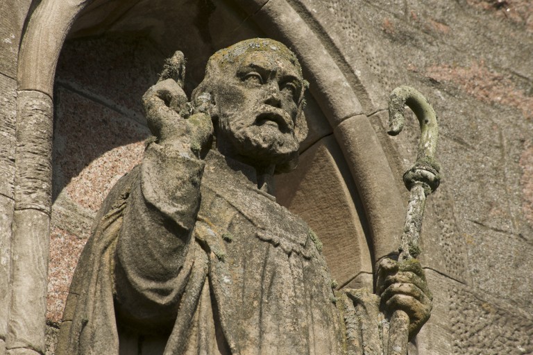 A stone depiction of St Colomba. He is bearded and carries a crooked staff in his right hand. His left hand is raised making a gesture. 