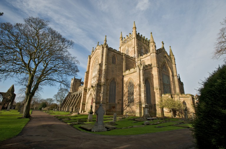 A general view of Dunfermline Abbey. A large tree, a path and gravestones are in the foreground. 