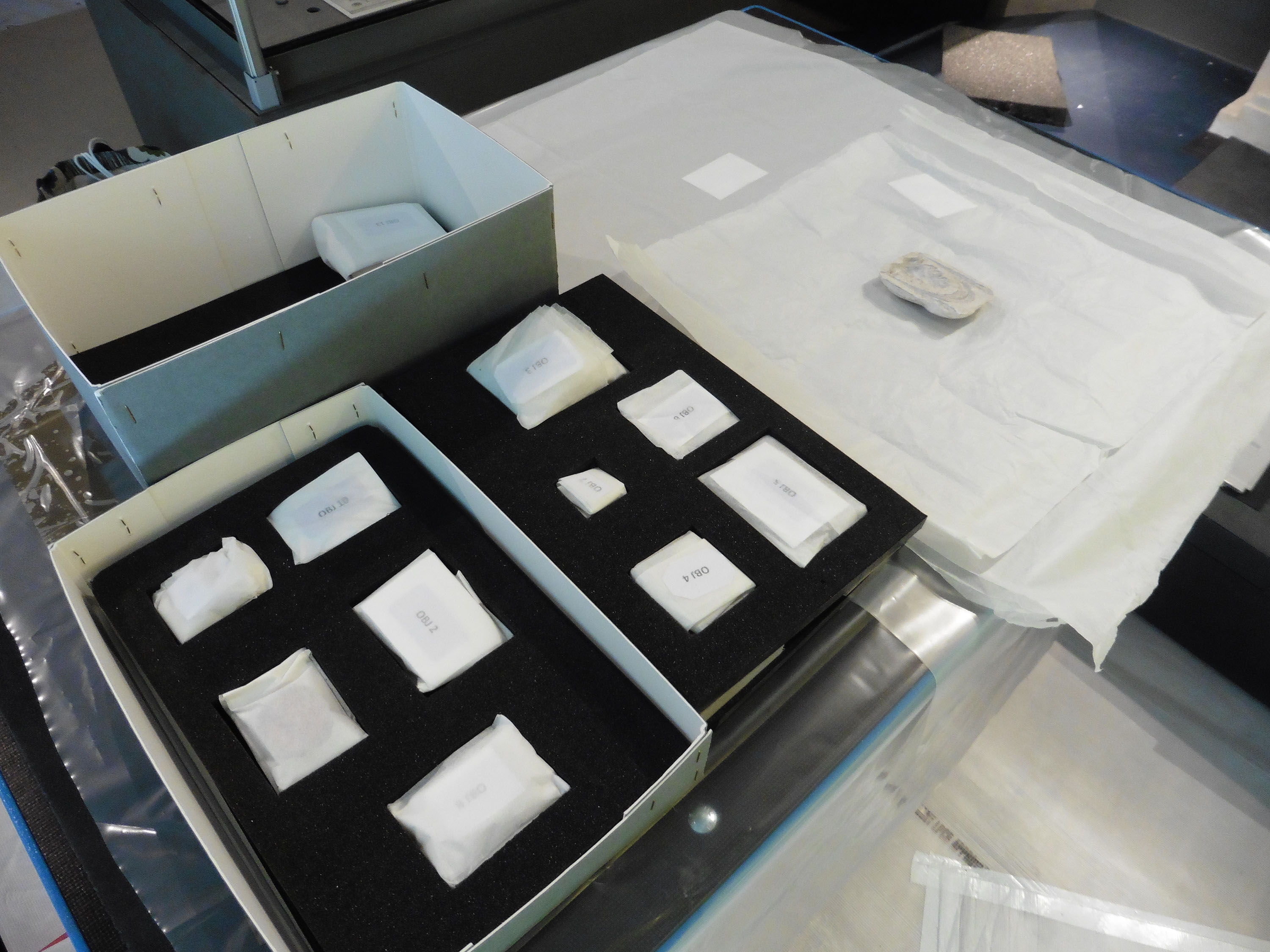 A box of objects is being unpacked on to a table at a museum. Each object is individually numbered and wrapped in white protective material.