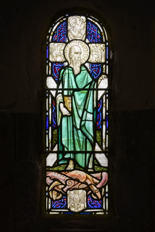 A stained glass window depicting St Andrew. He is dressed in green with a long white beard. In the background is a stone cross with a celtic design. 