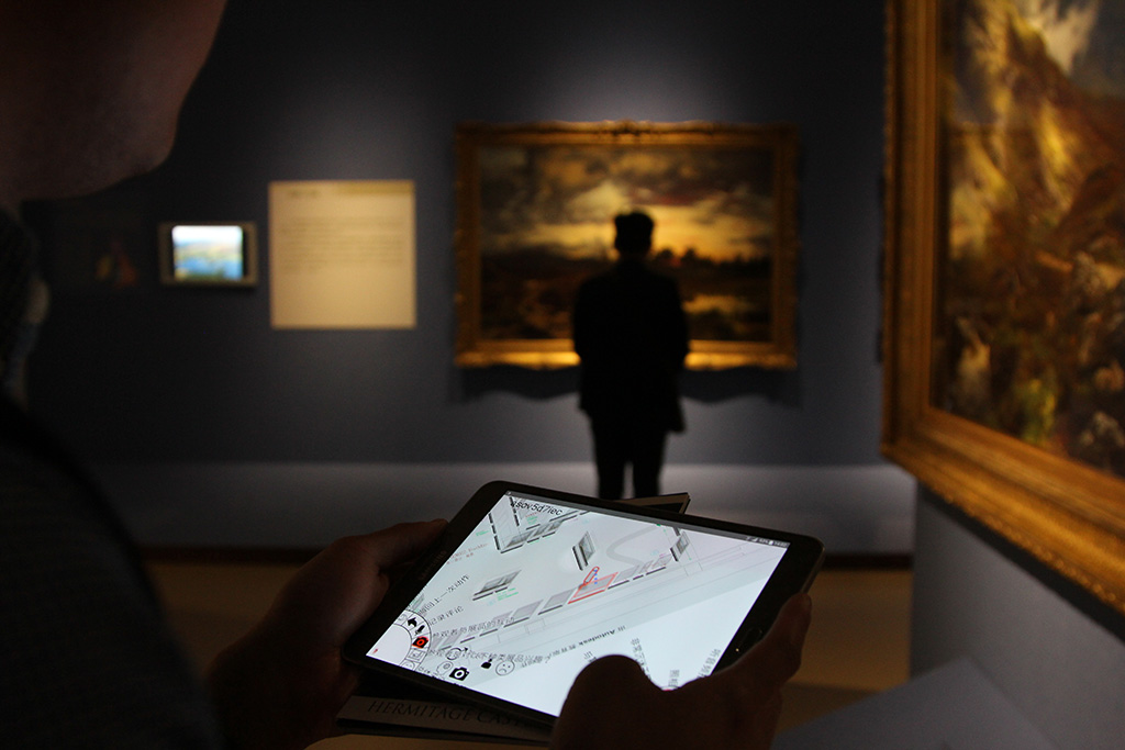 A mobile application is being used inside a museum exhibition . A man can be seen studying a framed picture on the wall. 