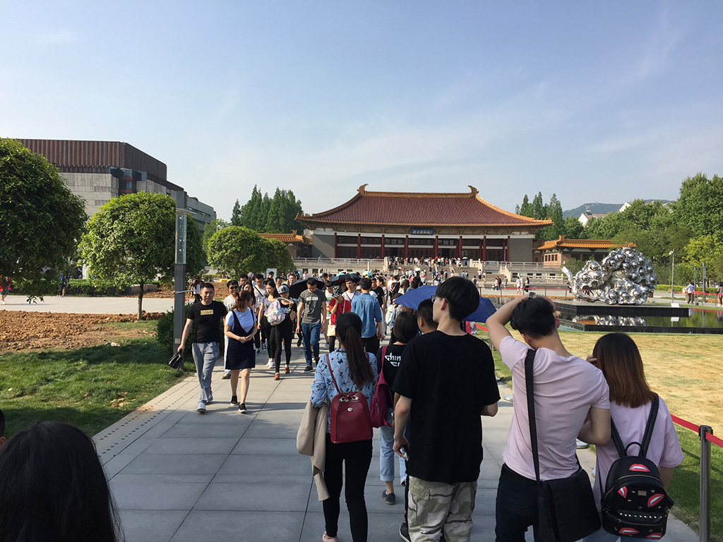 Visitors outside the Nanjing Museum in China. The museum has a distinctive red tiled roof. There is a silver sculpture and steps in front of the building. 