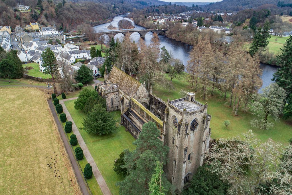 aerial view of Dunkeld Cathedral, a roofless church building lined with trees, with a river and bridge in the background