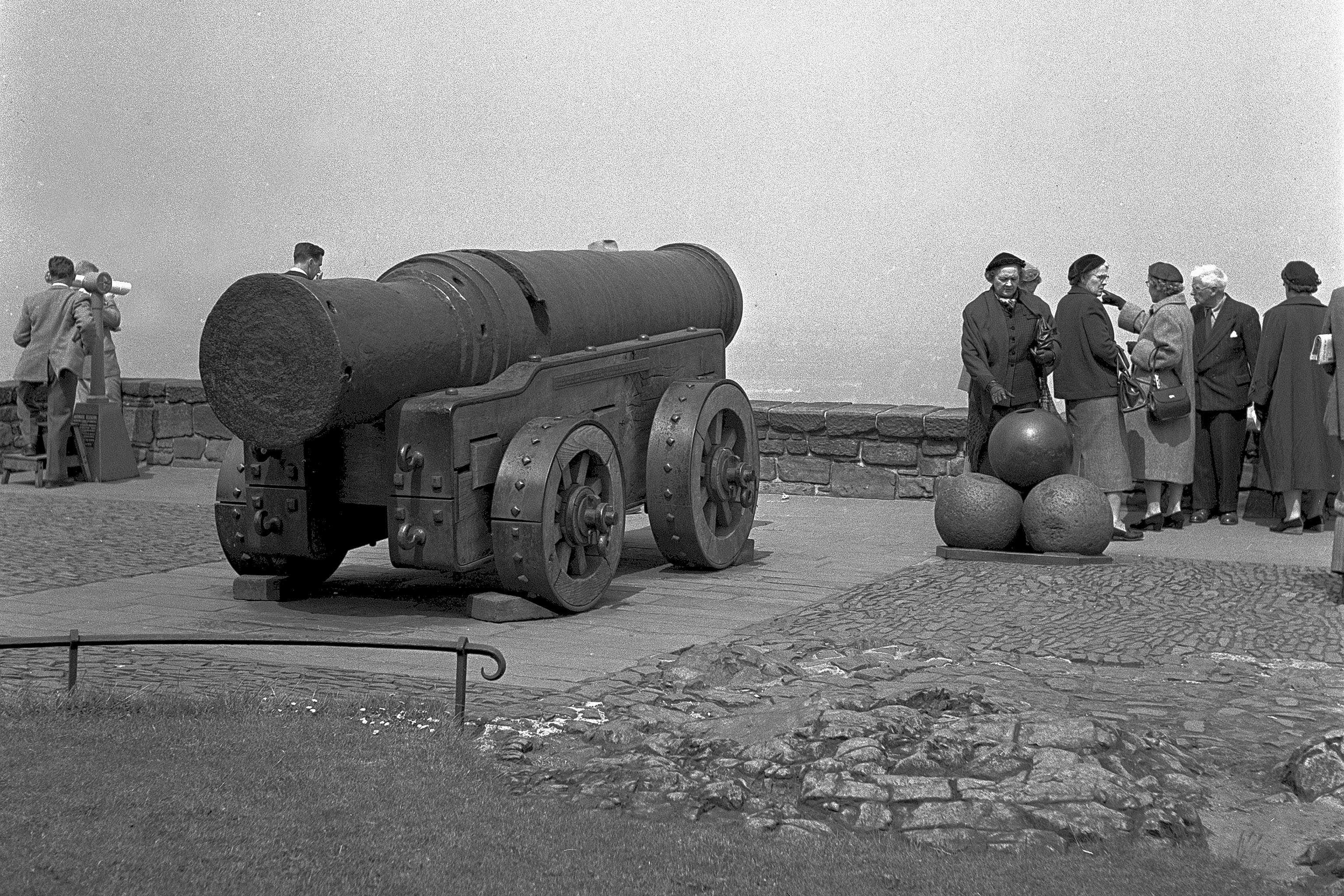 black and white image of an old cannon on wheeled carriage, pointed away from the camera as tourists stand to the right beside a pile of cannon balls