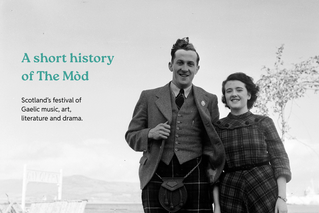 Black-and-white historic photograph of two people standing outdoors, one wearing a kilt and jacket and the other wearing a checked dress. On the left, teal text reads: “A short history of The Mòd” with the subheading, “Scotland’s festival of Gaelic music, art, literature and drama.”