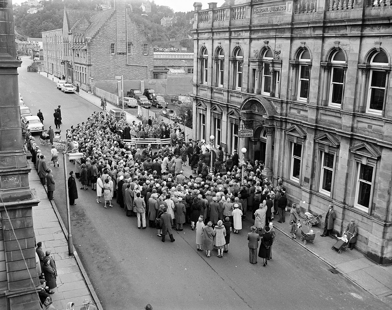 Black-and-white photograph of a large crowd gathered outside Oban’s municipal buildings during The Mòd. Viewed from above, hundreds of people fill the street and pavement around the entrance to the stone building, while parked cars, prams and surrounding town buildings can be seen in the background.