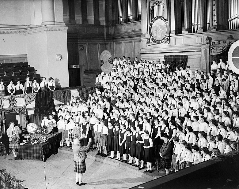 Black-and-white photograph of a large choir performing in Glasgow. Hundreds of singers stand on tiered risers inside a grand hall with wood-panelled walls and organ pipes, while a conductor in Highland dress stands at the front leading the performance.