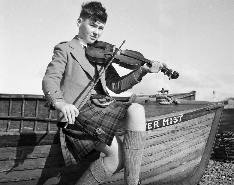 Black-and-white photograph of a young fiddler wearing a kilt and knee-high socks, seated on the bow of a wooden fishing boat. The musician holds a violin and bow while playing outdoors on a pebble beach, with the boat name “Silver Mist” visible on the hull.