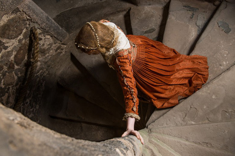 A model dressed as Mary Queen of Scots walks down a stone spiral staircase. She is wearing a red and gold dress.