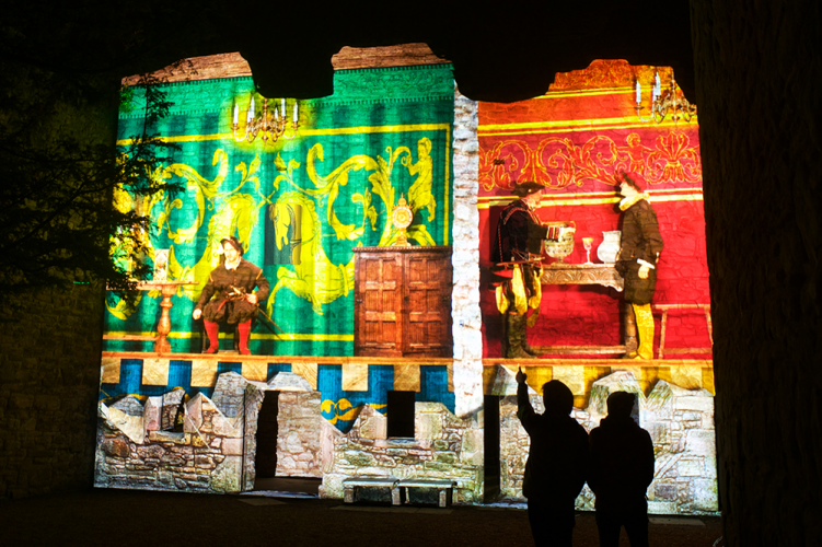 The silhouettes of two people watching a performance of Spotlight on Mary. The action is projected onto a castle wall. On the left hand side, a man with a sword sits at a table in front of a huge green tapestry featuring two gold unicorns. On the right, two men in 16th-century dress converse beneath a chandelier.