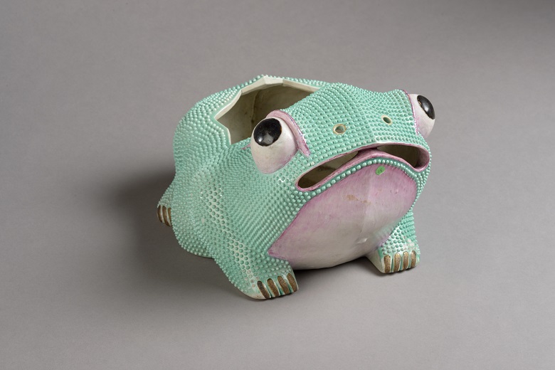 A green and pink ceramic flower holder in the shape of a frog. The frog is looking upwards with a bewildered expression on its face.