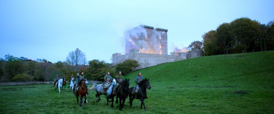 A still from the Outlaw King film. Nine medieval soldiers on horseback ride away from a burning castle.