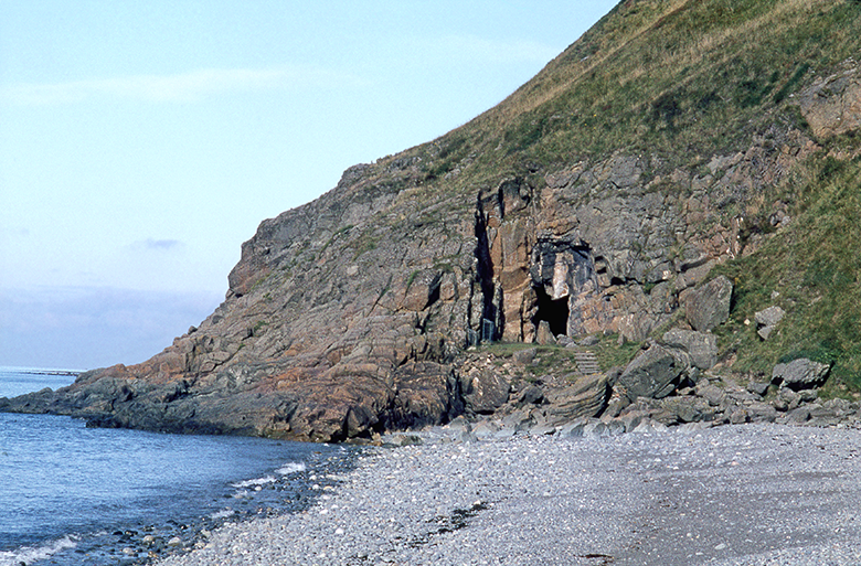 View across a pebble beach to a rocky outcrop where there is the mouth of a cave
