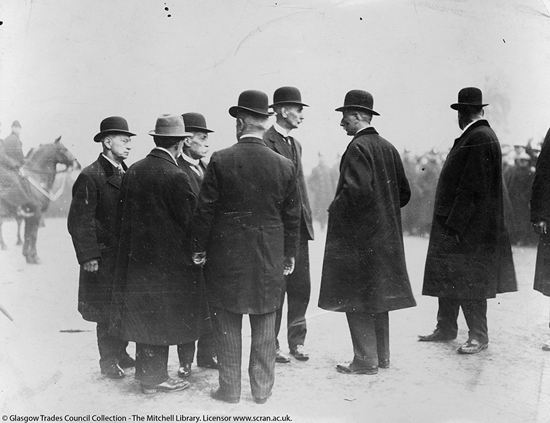 Eight men in coats and hats stand in discussion in front of a larger group of men. Mounted police can also be seen. 