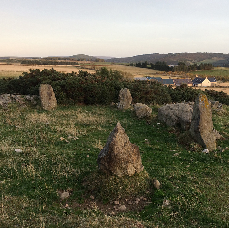 A close-up view of the Holmead stone circle with farm buildings and hills in the background.