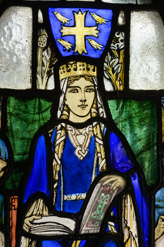 A stained glass depiction of St Margaret in St Margaret's Chapel, Edinburgh. She wears blue robes and a gold crown and has an open book , possibly a Bible, on her lap.