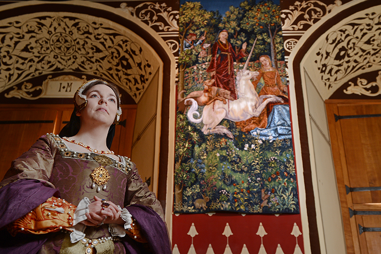 a woman dressed in renaissance clothes stands in front of the Hunt of the Unicorn tapestry, which shows the unicorn being attacked by hunting dogs