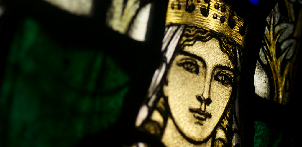 A close-up photo of a stained glass depiction of St Margaret. She has gold hair and wears a gold crown.