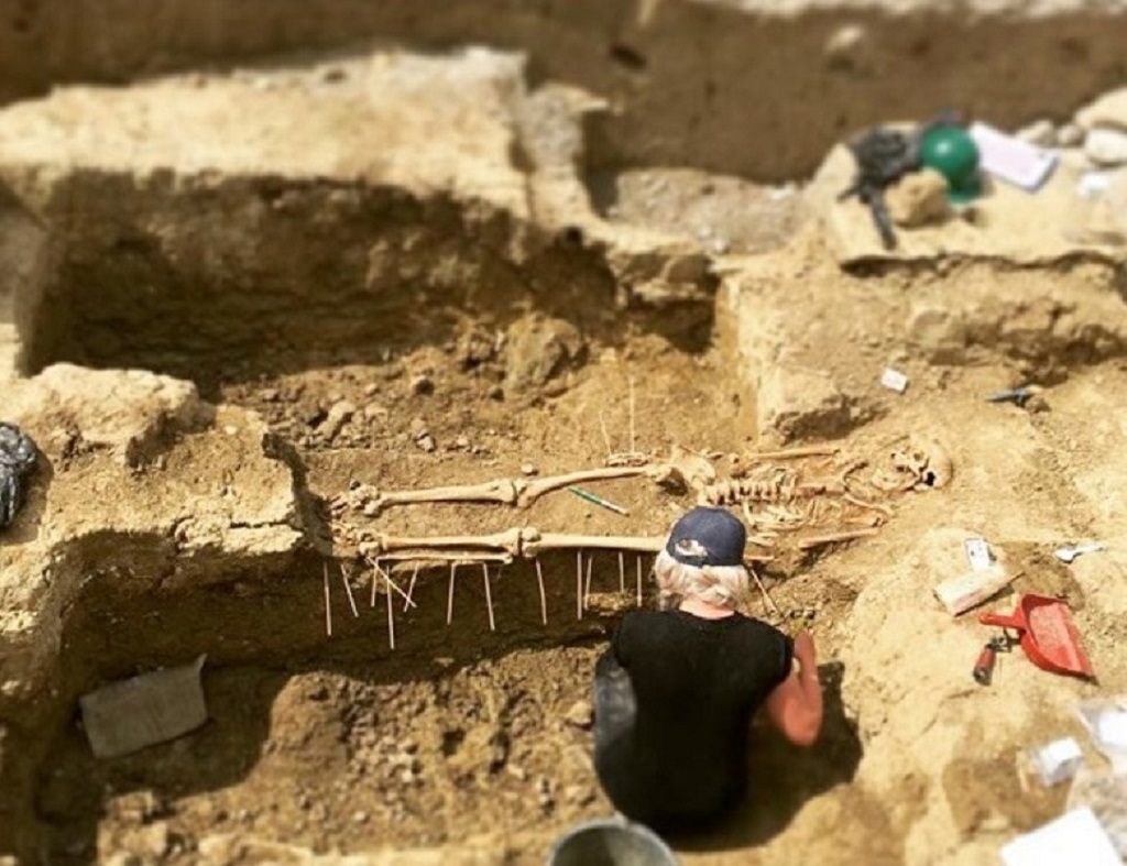An archaeologist working on site. She is crouched beside a human skeleton which has been uncovered.