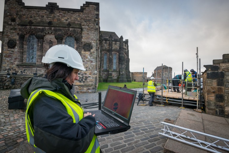 Lyn Wilson working with a laptop onsite at Stirling Castle. She wears a hard hat and reflective vest. 