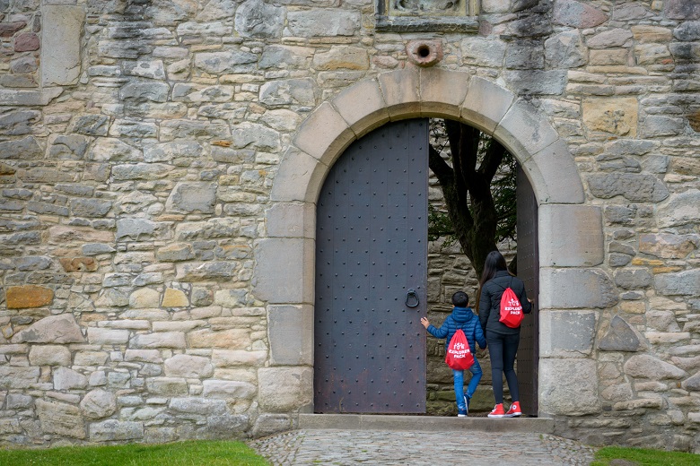Two young visitors opening the enormous front door of Craigmillar Castle. They are wearing red drawstring bags marked "Explorer Pack"
