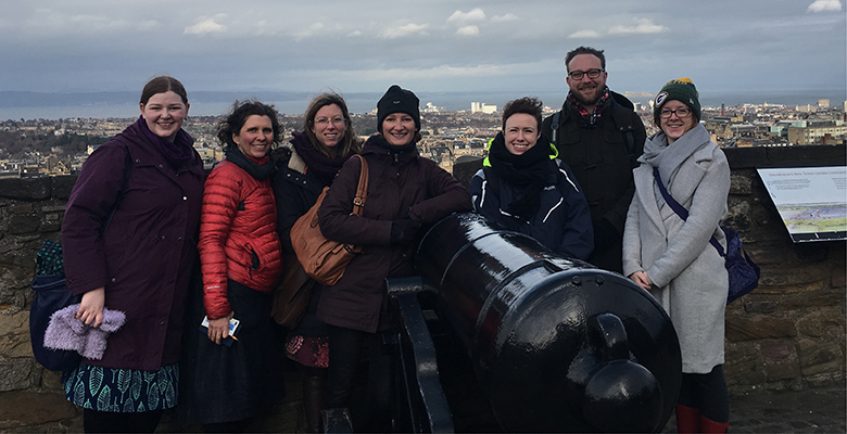 A group of seven people pose beside a large cannon on the battlements of Edinburgh Castle. The Firth of Forth can be seen in the background. 