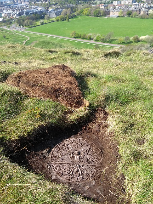 A brass plaque uncovered from beneath a layer of turf. Holyrood Park and some buildings of Edinburgh can be seen in the background.