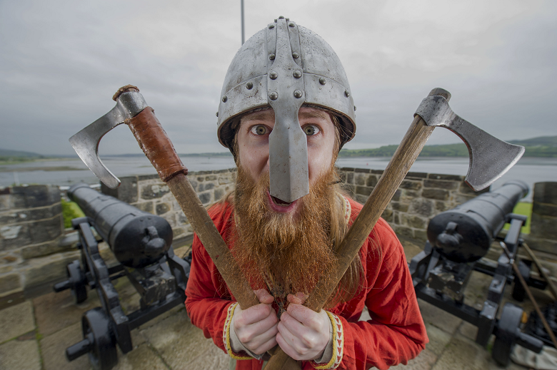 A bearded man wearing a metal helmet faces the camera with an axe in each hand 