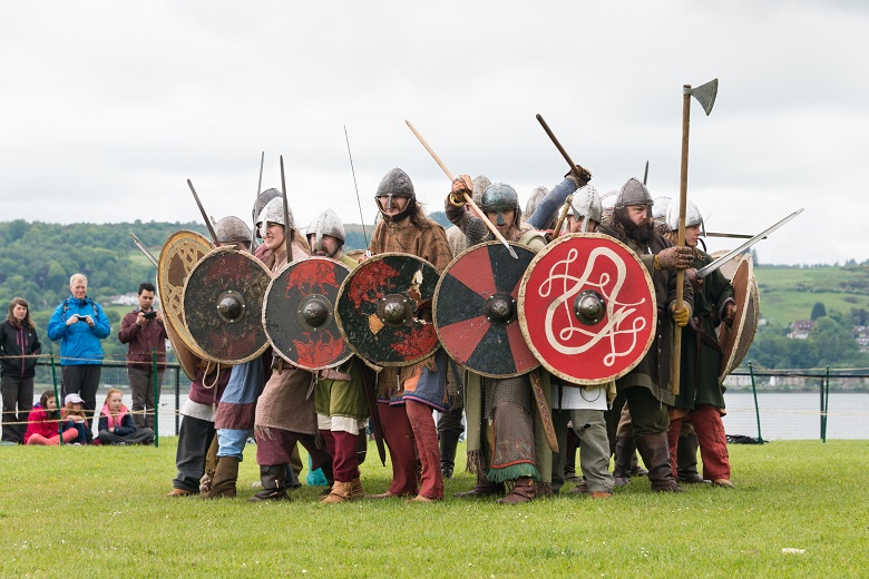 A group of reenactors dressed as Vikings in a battle formation. They are armed with swords, axes and colourful round shields. 