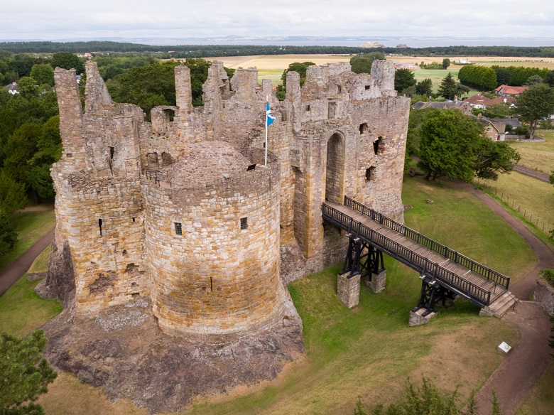 An aerial photo of Dirleton Castle showing the defensive might of the ruin, accessed by a long drawbridge over a dry moat. A saltire flies from the largest of the towers.
