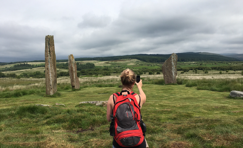 A lady wearing a rucksack takes a photo of three remote standing stones for Monument Monitor using a smartphone 