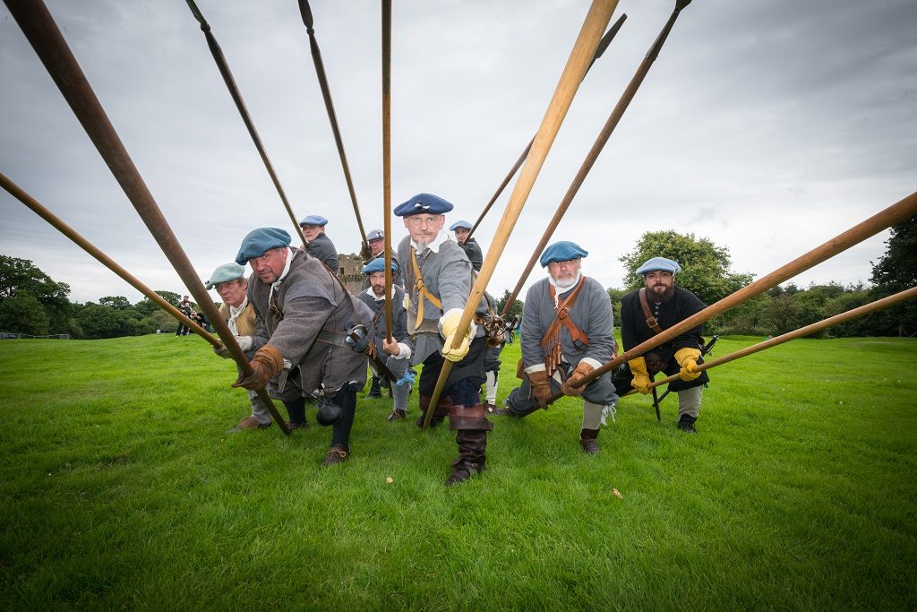 Around half a dozen men dressed as Covenanters (wearing tweed and blue flat caps). They brandish pikes at the camera.