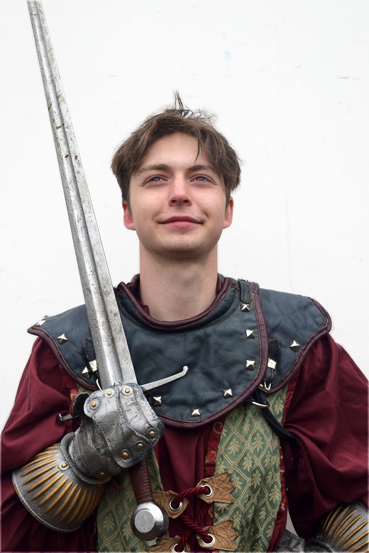 A young, confident jousting knight wearing a red and black outfit and holding a sword