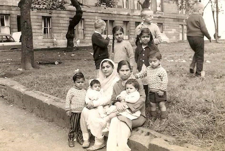 two women sit outside a tenement. Both hold babies. One wears a head scarf. They are surrounded by children.