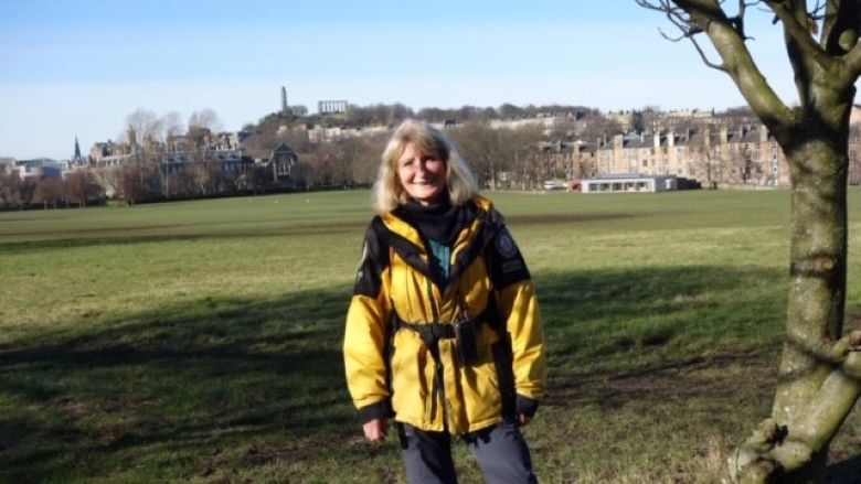 A lady wearing a black and yellow Ranger jacket stands in Holyrood Park with the education centre behind her