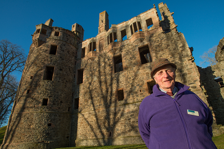 Patrick Scott, a volunteer guide at Huntly Castle, stands in front of the ruined castle in a purple sweater.