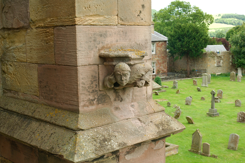 A stone carving of a man's face high on the wall of Melrose Abbey overlooking gravestones