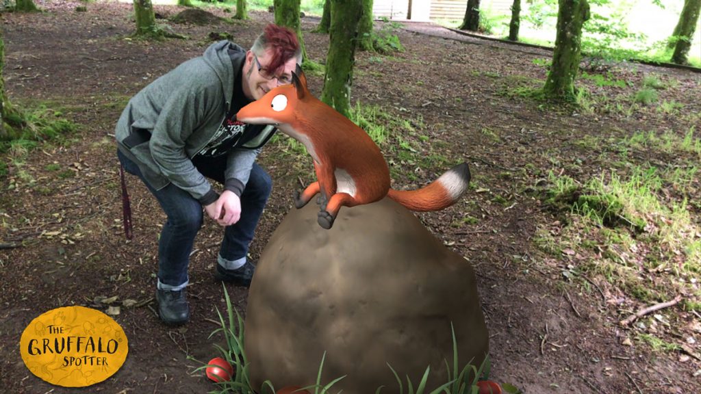 A man in a green hoodie and glasses smiling behind a cartoon fox sitting on a rock.