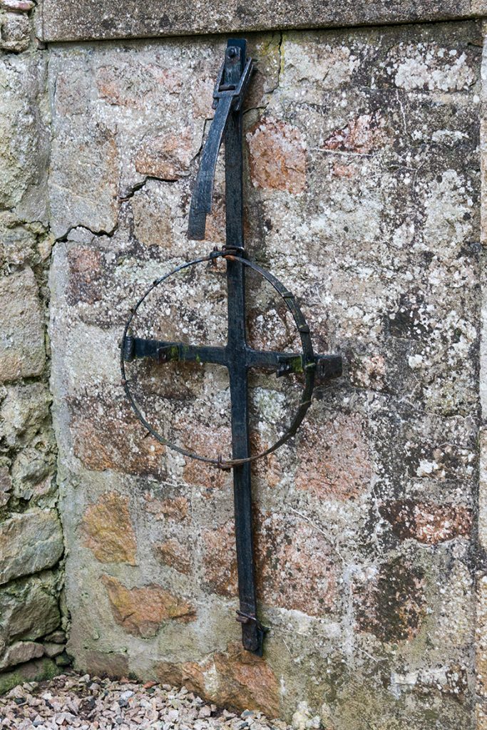 A large iron object hangs on a stone wall. There is a cross-shaped base and sitting on top of that are two semi-circular "jaws" with teeth.
