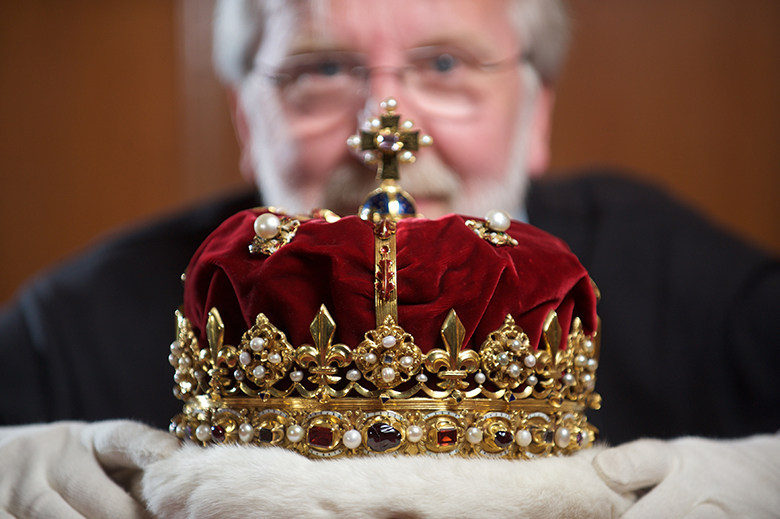 In the foreground is an ornate gold crown with red velvet fabric. Behind and out of focus is Richard Welander, the curator, who is holding the crown.
