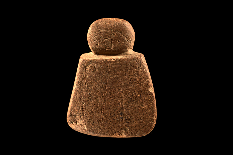 an abstract-looking carving. There is a round head and square body with minimal amounts of carved detail. On the head there are two small pin-hole eyes and a square for the nose. On the body there are two circles which have been interpreted as breasts, but could also be clasps for a cloak.
