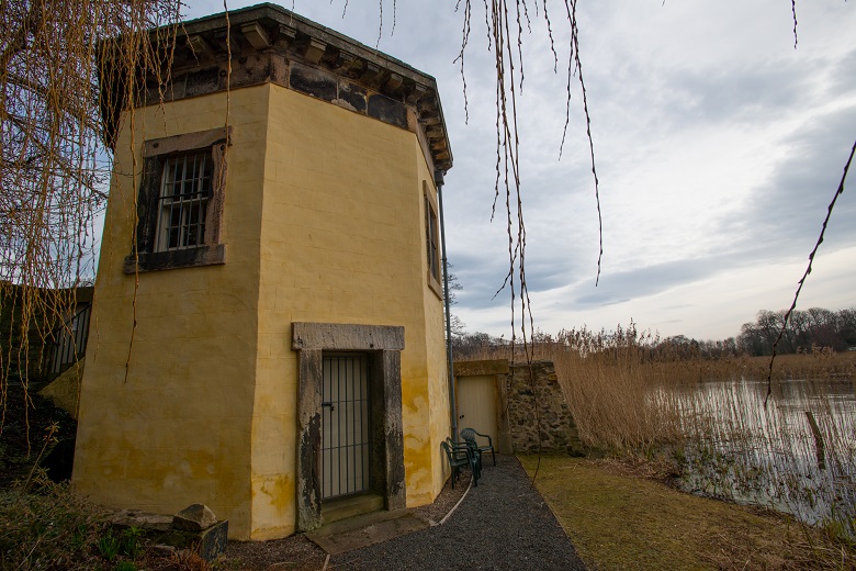 A hexagonal two storey tower close to the shore of a loch. It is painted yellow and is framed by the branches of a willow tree. 