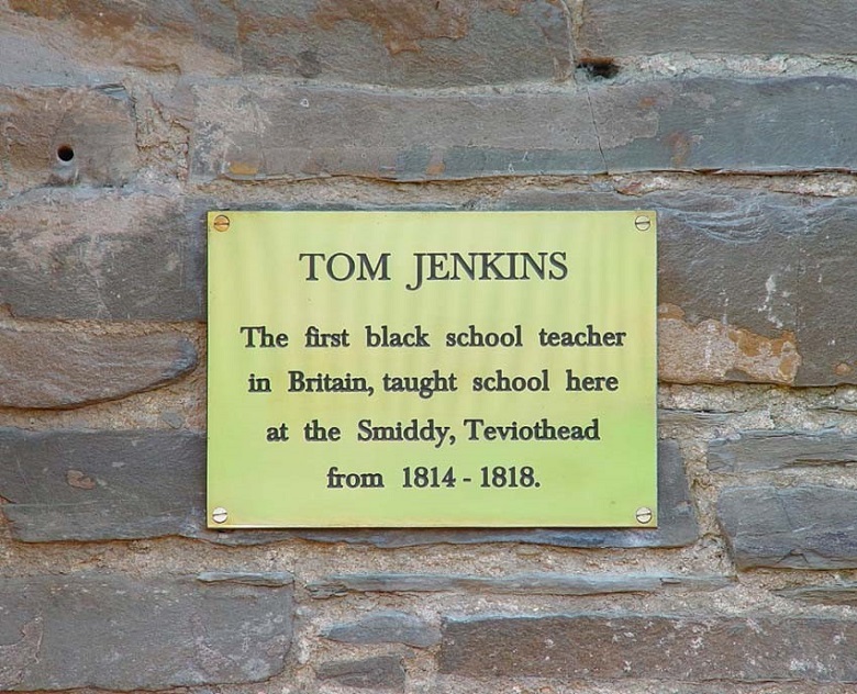 A small brass plaque on a brick wall. It reads "Tom jenkins. The first black school teacher in britain, taught schooll here at the Smiddy, Teviothead from 1814 - 1818." 